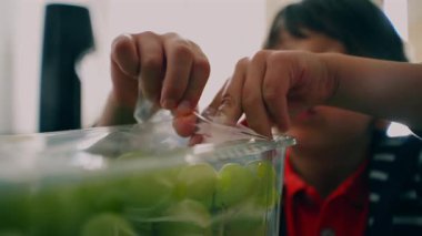 Genç çocuk dikkatle yeşil üzümlerden oluşan bir kabın içindeki plastik örtüyü soyuyor, odaklandığını ve evdeki basit, günlük aktivitelere odaklandığını vurguluyor..