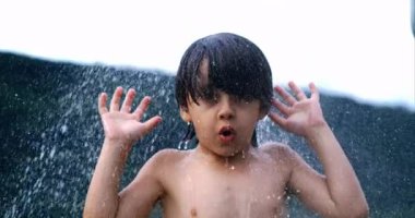 Genç bir çocuk yavaş çekimde serinletici bir su sıçraması yaşıyor. 800 fps hızla, elleri havada, açık havada, bulanık doğal bir geçmişi var.