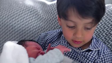 Küçük çocuk, yeni doğan kardeşine ilgi ve şefkat karışımıyla bakar. Bebeği dikkatlice kanepeye koyar ve kardeşler arasındaki koruyucu bağı sembolize eder..