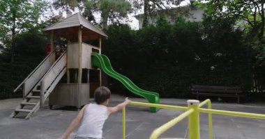 Parktaki sarı bir atlıkarıncada elinde sopa tutan genç bir çocuk, ahşap bir oyun evi ve arka planda yeşil yapraklarla çevrili bir kaydırak..