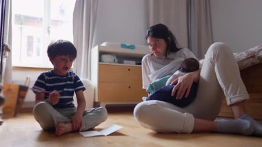 Anne, kucağında bebekle, bir ev okulu çizimi sırasında oğluna destek oluyor. Çok yönlü bir aile anı yakalıyor. Öğrenme, yaratıcılık ve beslenme..