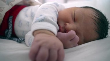 Yeni doğan bebek bir eli açık, diğer eli yüzü yakın bir şekilde bir yatakta huzur içinde uyuyor. Çocuğun sakin ifadesi ve rahat duruşu bir huzur ve güvenlik anını vurguluyor