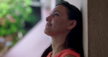 Bir kadın duvara yaslanıyor, sakin bir ifadeyle yukarı bakıyor, bulanık yeşil bir arka plana yakın çekim, yansıma ve huzur anı