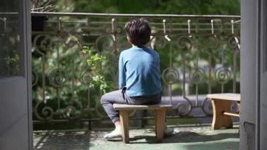 Genç bir çocuk balkon taburesinde sessizce oturuyor, yüzünü çevirmiş, dışarıda huzurlu bir anın tadını çıkarıyor. Huzurlu bir çevreye yansıyan bir çocuğun yalnızlığı ve sakinliği.