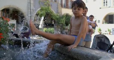 Küçük bir çocuk büyük sıçramalarla dolu bir çeşmede su tekmeliyor, 800 fps yavaş çekimde eğlenirken annesi ve küçük kardeşi arka planda izliyor..