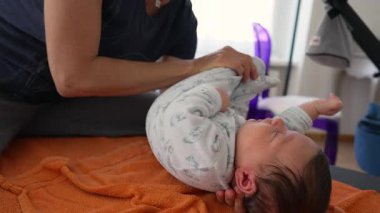 Ortopedist, çocukluk yıllarında vücut ağrılarını hafifletmek için turuncu bir havluyla yeni doğmuş bebeklerin bacaklarını esnetiyor. Bebek rahat bir tulum giyer, nazik ve profesyonel bakım görür.
