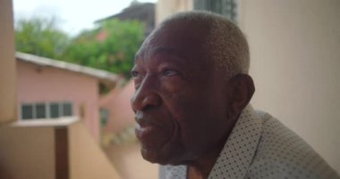 Yaşlı siyah bir adam verandada bir sandalyede oturuyor, sakin bir ifadeyle mesafeye bakıyor, tropik bir ortam, pembe bir bina, arka plandaki merdivenler.