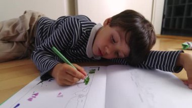 Yerde yatarken yeşil bir kalemle çizim yapan genç bir çocuk, bir kitaptaki boyamaya odaklanmış, yoğun yaratıcı konsantrasyon, kapalı alanda sanat zamanı.