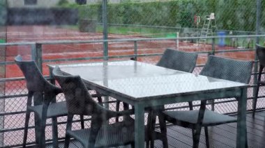 Yağmurlu bir günde dışarıda yemek masası ve sandalyeler, gri gökyüzünü yansıtan ıslak yüzeyler, çitli bir spor salonuna bakan, sessiz ve boş bir sahne.