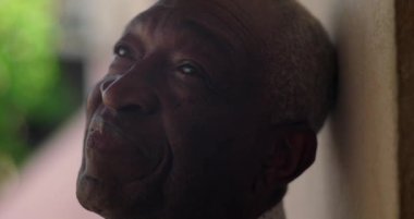 Afrika kökenli yaşlı bir adam duvara yaslanmış, sakin ve sakin bir ifadeyle yukarıya bakıyor, açık hava, canlı arka plan, yansıtıcı ve barışçıl