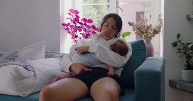 Anne bebeğini kucağında, bebek yüzüne dokunurken yavaşça kanepede dinleniyor. Huzurlu bir ev ortamında samimi bir yakınlık ve bağ anı.