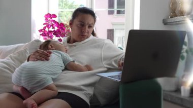 Anne kucağında bebek uyurken dizüstü bilgisayar kullanıyor, iş ve çocuk bakımını yönetiyor, uzak işleri dengeliyor ve ev ortamında ebeveynlik sorumluluklarını dengeliyor