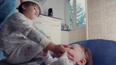 Pijamalı bir çocuk, rahat bir mutfakta küçük bir kardeşle şakalaşıyor. Sıcak, kapalı bir ortamda sevgi ve ilgi dolu bir kardeş anı..