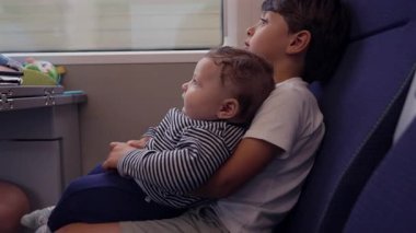 Kardeşini trende tutan büyük kardeş, ikisi de merakla yukarı bakıyor, çocukluk bağı kurma anı, paylaşılan seyahat deneyimi, toplu taşıma, rahat ve güvenli.