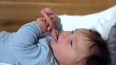 Bebek yumuşak yatakta uzanıyor. Kirpi tasarımı ile diş çıkarma halkası çiğniyor. İki eliyle oyuncağı kavrıyor.
