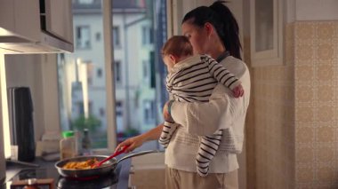 Anne mutfakta yemek pişirirken bebeği kucağına alır, tavada yemek karıştırır, kucağında çocukla birden fazla iş yapar, evde ebeveynlik görevleri yapar.