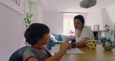 Anne, oğluyla evde eğitim materyalleri ve ev eşyalarıyla dolu bir masada, parlak, modern bir oturma odasında otururken hayal kırıklığını ifade ediyor.