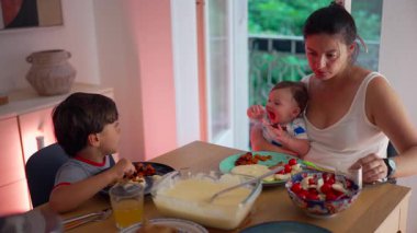 Anne yemek masasında bebeği tutuyor, büyük çocuk yemek yerken onu rahatlatıyor, bakıcılık ve yemek zamanını dengeliyor, bakım ortamı, aile birliği, ev yapımı akşam yemeği.