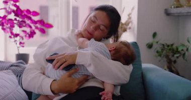 Anne bebeğini kanepede tutuyor, yüzü hem yorgunluk hem de sakinlik gösteriyor, anne olmanın günlük gerçekliğini yansıtıyor, rahat ve huzurlu bir yuvada.