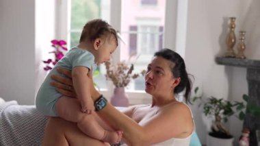 Anne kucağında bebek, sevgi dolu bakışlar, neşeli bir bağ, neşeli bir etkileşim, anne gülümseyişi, bebek aşağı bakıyor, ebeveyn-çocuk bağı, sıcaklık ve evde sevgi.