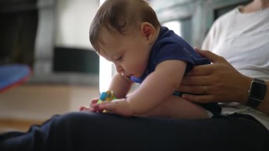 Bebek anne kucağında öne eğiliyor, oyuncağa uzanıyor, odaklanmış keşif, erken gelişim, duyusal öğrenme, merak, bakım, destekleyici ebeveynlik