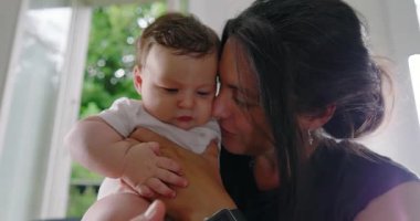 Anne sevgiyle bebeğini kucağında tutuyor, alınları birbirine değiyor, samimi ve duygusal bir bağ kurma anını paylaşıyor, sıcak bir yuva ortamında derin bir sevgi ve bağlantı kuruyor.