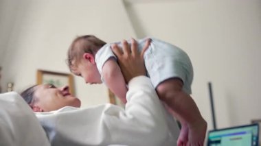 Anne bebeği havaya kaldırıyor, neşeli yüz ifadesi, neşeli bağlanma anı, bebek gülüşü, eğlenceli etkileşim, mutlu aile zamanı, ebeveyn-çocuk bağı, enerjik hareket