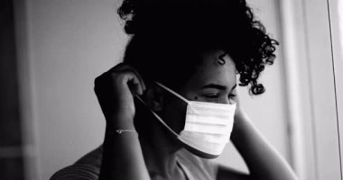 Genç siyahi bir kadın, yoğun dramatik monokromatik portresiyle evde takılıp kalan siyah maskesini takıyor. Daireden pencereye bakıyor.