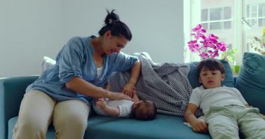 Annesi kanepede oturup bebeğine özenle bakarken büyük oğlu rahat bir poz veriyor. Rahat, doğal ışıklandırılmış bir oturma odasında oturuyor.