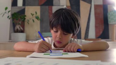 Genç bir çocuk, mavi bir kalemle bir meydanı boyuyor, bir masada yaratıcı aktivitelere odaklanıyor, konsantrasyon ve sanatsal gelişim gösteriyor..