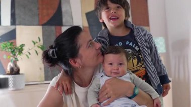 Anne bebeğini kucağına alıp öperken, büyük kardeş gülümseyerek izliyor. Evde anne, bebek ve kardeşle iç içe geçen iç açıcı bir aile anı..