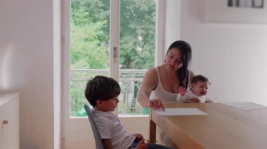 Anne evde eğitim ve çocuk bakımını dengeliyor, bir bebeği kucağında tutarken oğluna okul ödevlerinde yardım ediyor, evde öğrenim ortamında sabır ve destek oluyor.