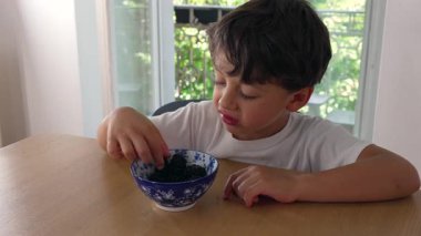 Genç çocuk masada oturmuş böğürtlen yiyor, sağlıklı atıştırmalıkların tadını çıkarıyor, böğürtlen dolu bir kase, doğal gün ışığı, ev dekorasyonu, rahatlama anı, çocukluk beslenmesi.