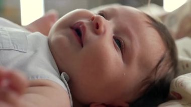 Bebek yatakta uzanıyor, belli belirsiz bir gülümsemeyle yukarı bakıyor. Yakından bakıldığında bebeğin yanakları ve yumuşak saçları vurgulanıyor. Bebek ise rahat ve rahat bir ortamda.