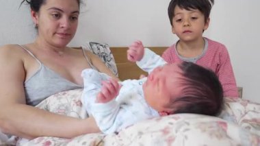 Anne ve büyük kardeş bebekle yatakta oynuyor, nazik etkileşim, hassas bağ anı, şefkatli dokunuş, kardeş bağı, rahat yuva kurma, sıcak aile ortamı.