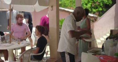 Dışarıdaki aile toplantısı sırasında kendisine yemek servisi yapan yaşlı bir adam, çeşitli insanlarla çevrili sıcak ve misafirperver bir atmosferde gündelik bir yemeğin tadını çıkarıyor.