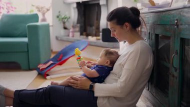 Anne kucağında bebekle oturuyor, oyuncaklarla oynuyor, meşgul ve odaklanmış, bağlanma anı, erken çocukluk gelişimi, ebeveynlik, sıcak ve sevgi dolu bir ev.
