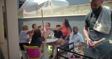 Bir grup farklı arkadaş ve aile bardaklarını açık hava barbeküsünde kaldırıyor, büyük bir şemsiye altında bir masanın etrafında birlik ve neşeyi canlı bir arka bahçede kutluyorlar.