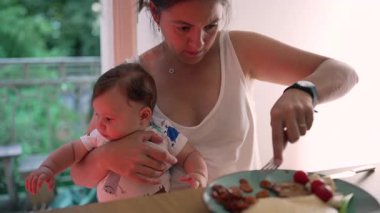 Anne kucağında bebek varken yemek kesiyor, çoklu görevlere odaklanıyor, çocuk bakımı ve yemek zamanını dengeliyor, günlük ebeveynlik, gerçek hayatta annelik, evde yemek yeme deneyimi