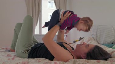 Anne yatağa uzanıp bebeği iki eliyle büyütüyor, neşeli bir bağ kuruyor, aile yatak odasında ebeveyn-çocuk ilişkisini güçlendiriyor.