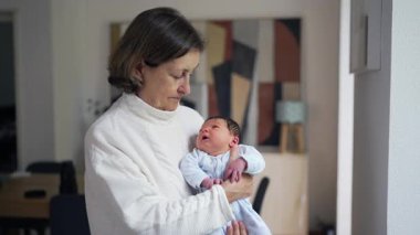 Yeni doğmuş torununu kucağında tutan yaşlı bir kadın sıcak, sevgi ve şefkatli bir bağ taşıyor, rahat, doğal ışıklandırılmış bir oturma odasında.