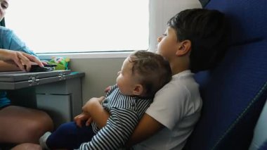 Büyük kardeş, bebek kardeşini tren koltuğunda tutuyor, pencereden dışarı bakıyor, aile bağı, çocukluk merakı, şefkatli kucaklama, ortak seyahat tecrübesi, sessiz bir yolculuk.