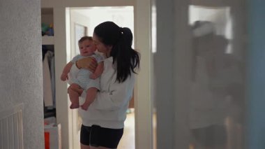 Anne bebeğini koridorda kucağında tutuyor. Kapının önünde bebeğini yanağından öpüyor. Yumuşak ışıklandırma bağlarını vurgular.