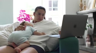 Anne kucağında uyuyan bebekle evden çalışıyor, uzaktan kumandalı işi ve ebeveynliği dengeliyor, rahat oturma odasında çocukla ilgilenirken laptopta birden fazla iş yapıyor..