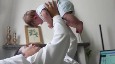 Anne bebeği havaya kaldırıyor, bebek neşeli bir ifadeyle gülüyor, neşeli bir bağ, eğlenceli etkileşim, ebeveyn-çocuk bağı, enerjik an, mutlu aile zamanı, kaygısız neşe.