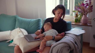 Anne kucağında bebekle dizüstü bilgisayarda çalışıyor, ebeveynlik ve işle uğraşıyor, ev ofisi, modern aile hayatı, uzak iş, kariyer dengesi, çalışırken bebeğe bakıyor..
