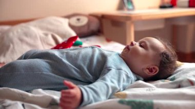Yatağa uzanmış bebek sakince etrafına bakıyor, açık kollu jest, gevşek ifade, rahat ve huzurlu yatak odası ortamı, duyusal farkındalığın erken aşamaları.