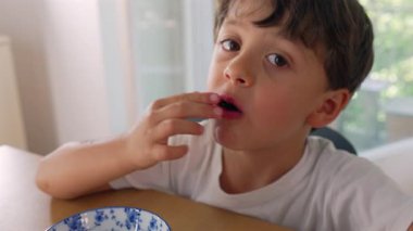 Masada böğürtlen yiyen genç bir çocuk, taze böğürtlen yiyor, meyve dolu bir kase, sessiz bir an, sağlıklı atıştırmalıklar, doğal gün ışığı, ev dekorasyonu, çocukluk beslenmesi..