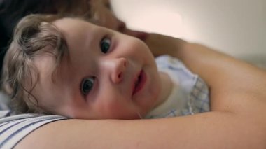 Annenin kollarında uzanan bebeğe yakın çekim, meraklı, rahat yüz ifadesi, şefkatli kucaklama, rahatlık, güven, sevgi dolu bağ, aile sıcaklığı..