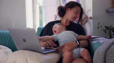 Anne kucağında bebek varken dizüstü bilgisayarda çalışıyor. Çoklu görevdeki ebeveyn, iş ve çocuk bakımını dengelemek, uzaktan çalışma, aile hayatı, modern annelik, ev işi..